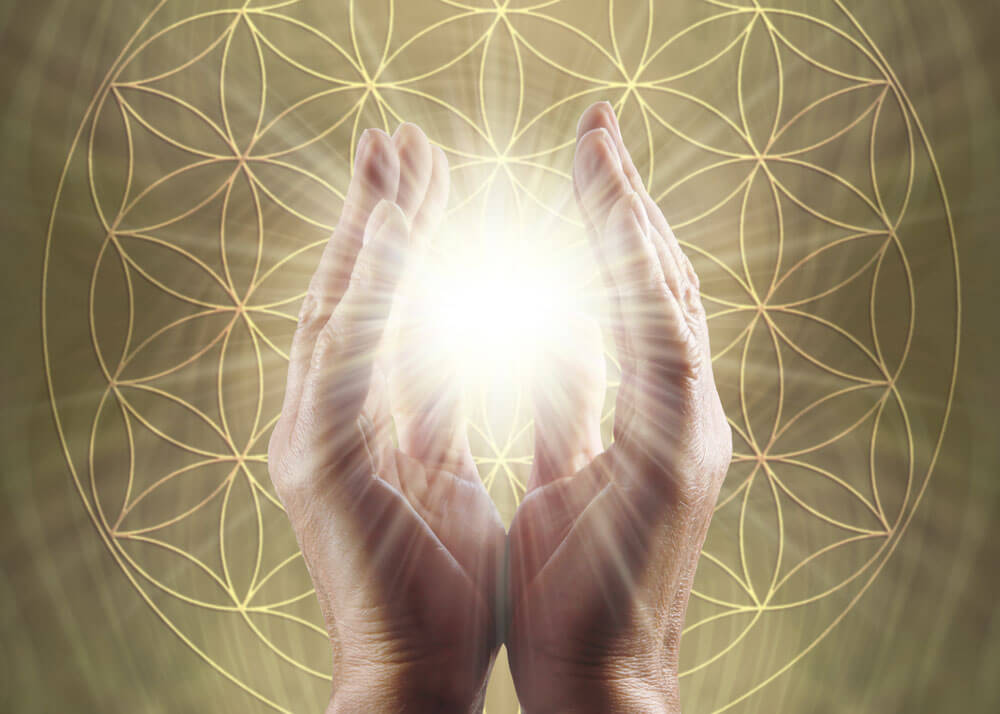 Two hands with palms facing each other and a glow of light shining bright between them with the flower of life symbol in teh background.